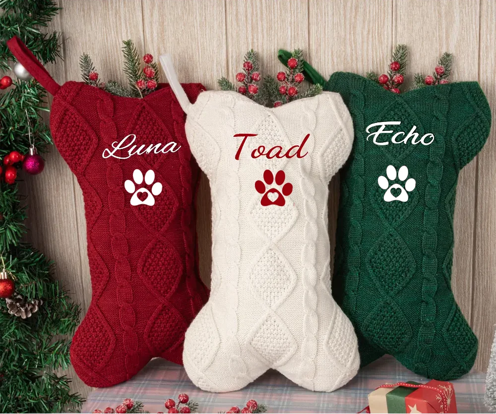 Three knitted dog xmas stockings in red, white, and green with names and paw prints on a wooden background.