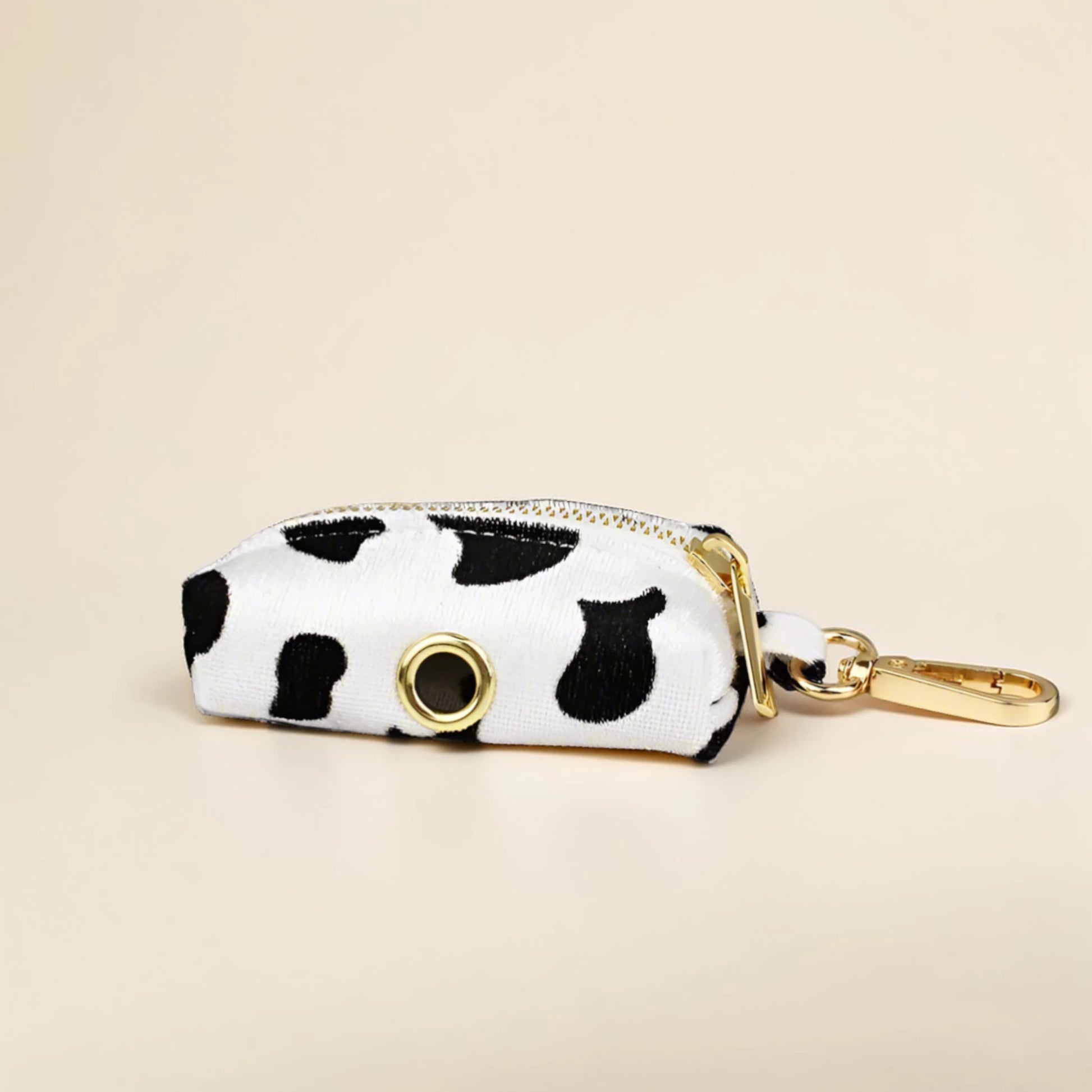 Cow print dog poop bag holder with gold zip and clip on a beige background