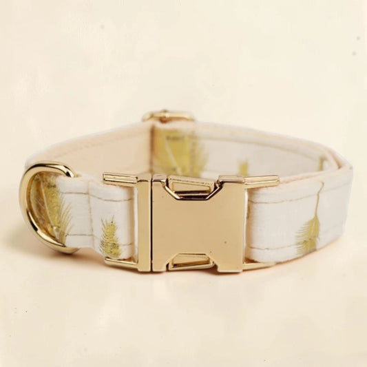 A dog collar with a cream-colored fabric strap featuring a subtle gold feather pattern, finished with a polished gold-tone metal buckle and D-ring.