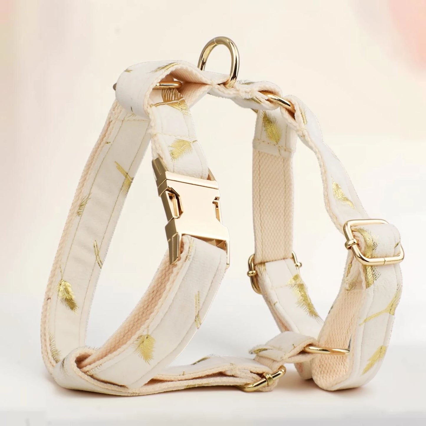 A cream-colored dog harness with a subtle gold leaf pattern, featuring gold-tone metal hardware, including a quick-release buckle, adjustment slider, and D-ring, set against a soft, light beige background.