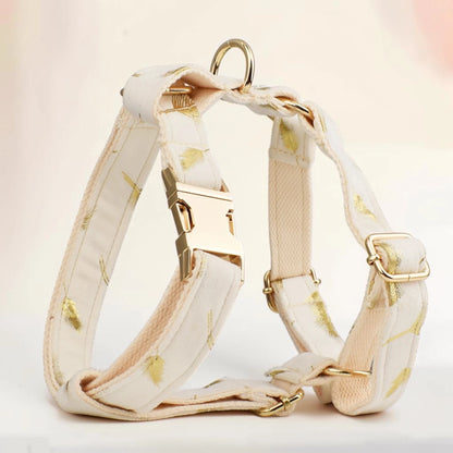 A cream-colored dog harness with a subtle gold leaf pattern, featuring gold-tone metal hardware, including a quick-release buckle, adjustment slider, and D-ring, set against a soft, light beige background.