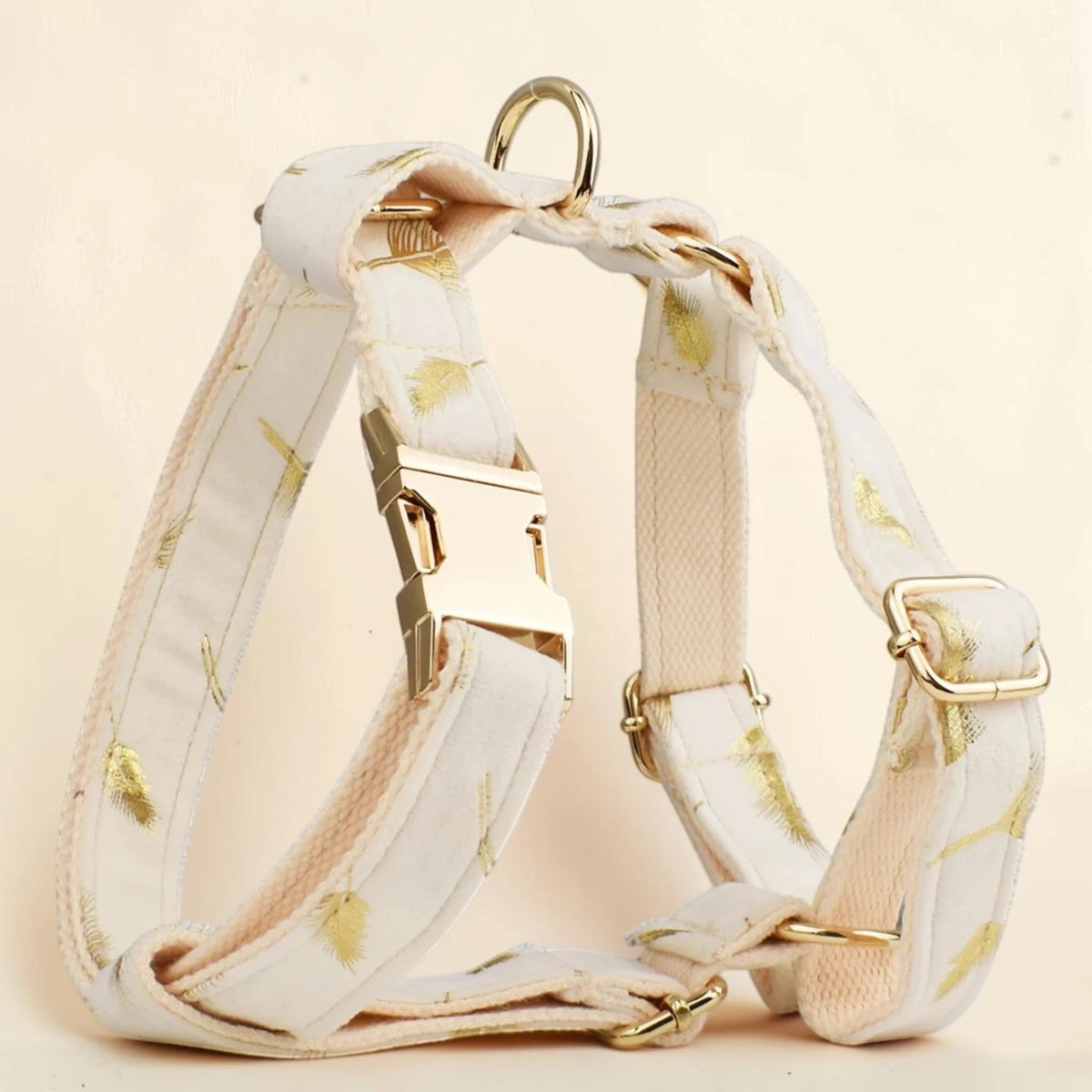 A cream-colored dog harness with a subtle gold leaf pattern, featuring gold-tone metal hardware, including a quick-release buckle, adjustment slider, and D-ring, set against a soft, light beige background.