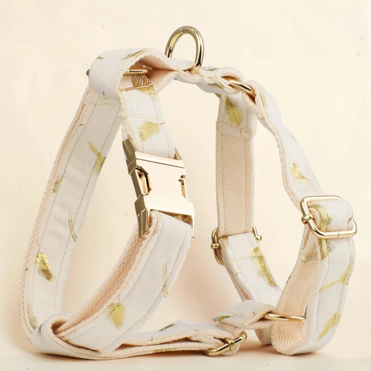 A cream-colored dog harness with a subtle gold leaf pattern, featuring gold-tone metal hardware, including a quick-release buckle, adjustment slider, and D-ring, set against a soft, light beige background.
