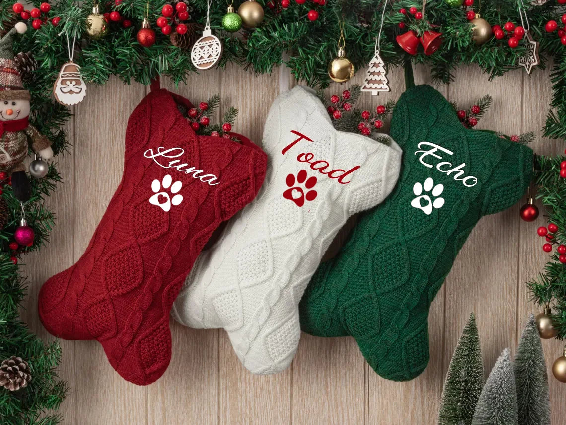 Three knitted pet xmas stockings in red, white, and green with names 'Luna', 'Toad', and 'Echo' on a wooden background with Christmas decorations.