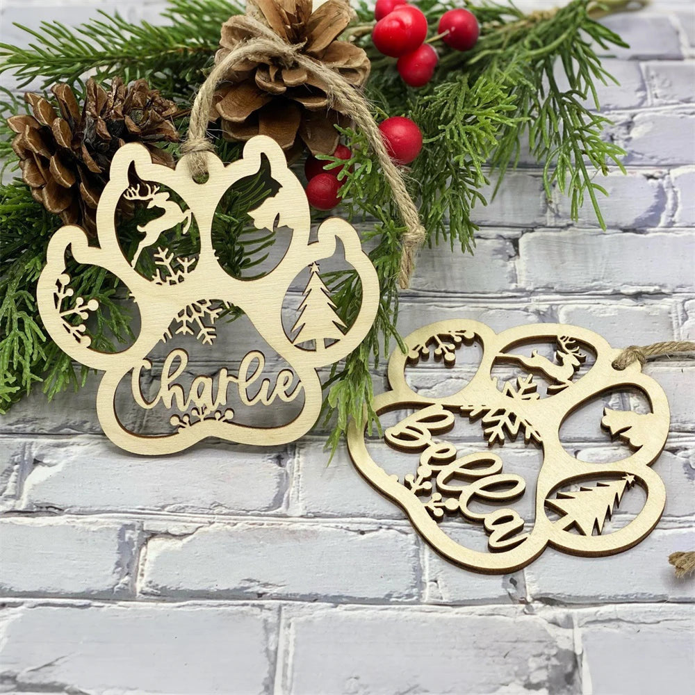 Two wooden paw-shaped ornaments with names 'Charlie' and 'Bella' on a decorative background with greenery and pinecones.